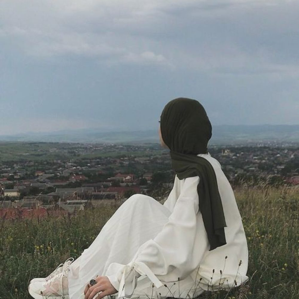 A female in white gown and dark green scarf sit on grass to enjoy with nature view
